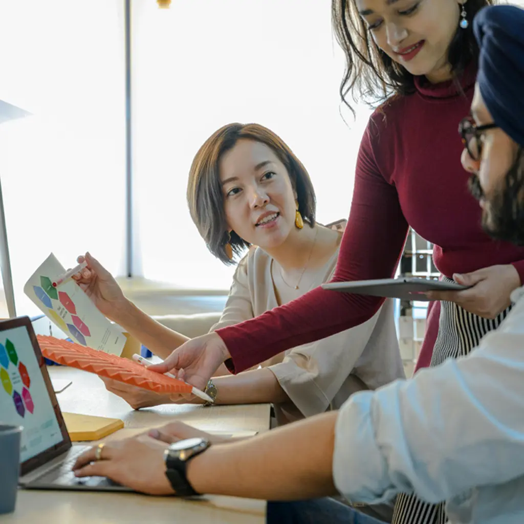 Three colleagues collaborate in an office, discussing colorful charts and a presentation. The mood is focused and collaborative, with technology in use.
